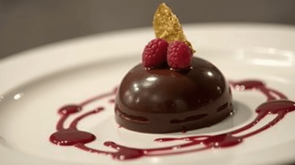 A close-up of a fancy dessert plated with a glossy chocolate dome, raspberry coulis, and edible gold leaf garnish.
