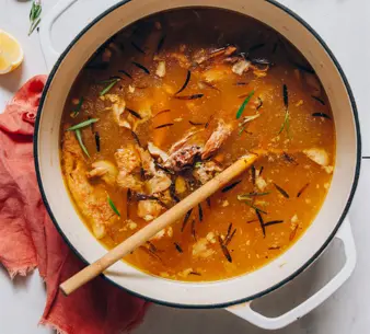 Chicken bone broth simmering in a white pot with herbs and chicken bones.
