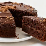 A moist chocolate cake on a white plate topped with glossy frosting and a slice pulled out, showing its soft crumb and rich texture.